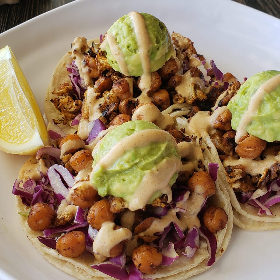 Three tacos topped with roasted cauliflower, chickpeas, purple cabbage, avocado, creamy sauce, and a lemon wedge on a white plate.