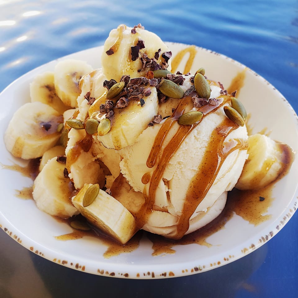 Banana-topped ice cream drizzled with caramel and sprinkled with cacao nibs and pumpkin seeds on a white plate.