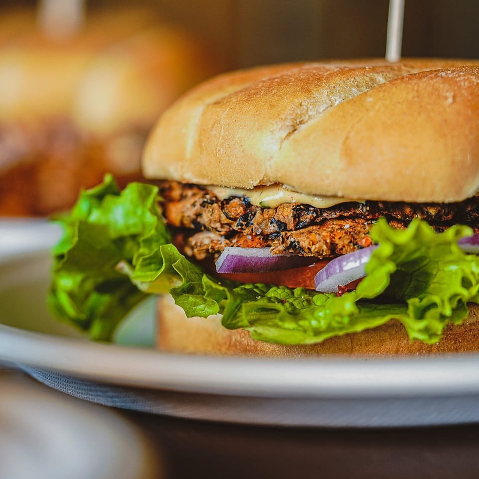 Close-up of a hearty burger with a toasted bun, grilled patty, lettuce, tomato, red onion, and sauce on a plate.