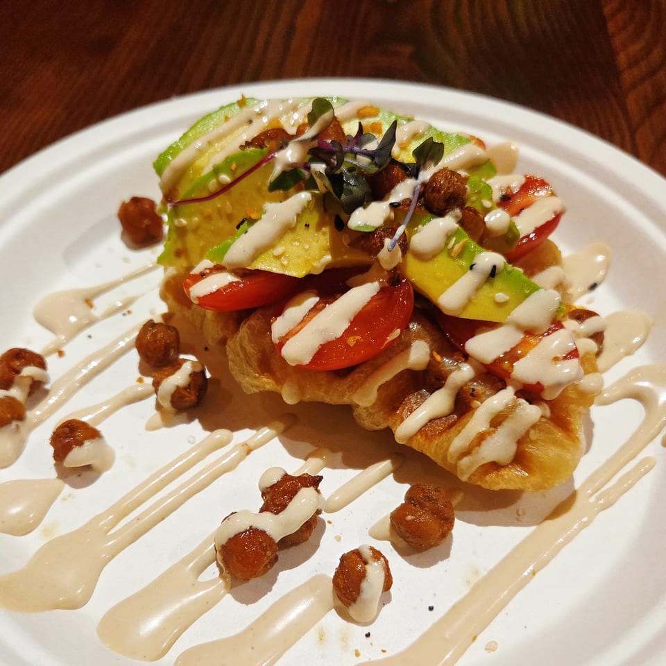 Crispy pastry topped with avocado, tomatoes, greens, crunchy chickpeas, and creamy white sauce on a white plate.