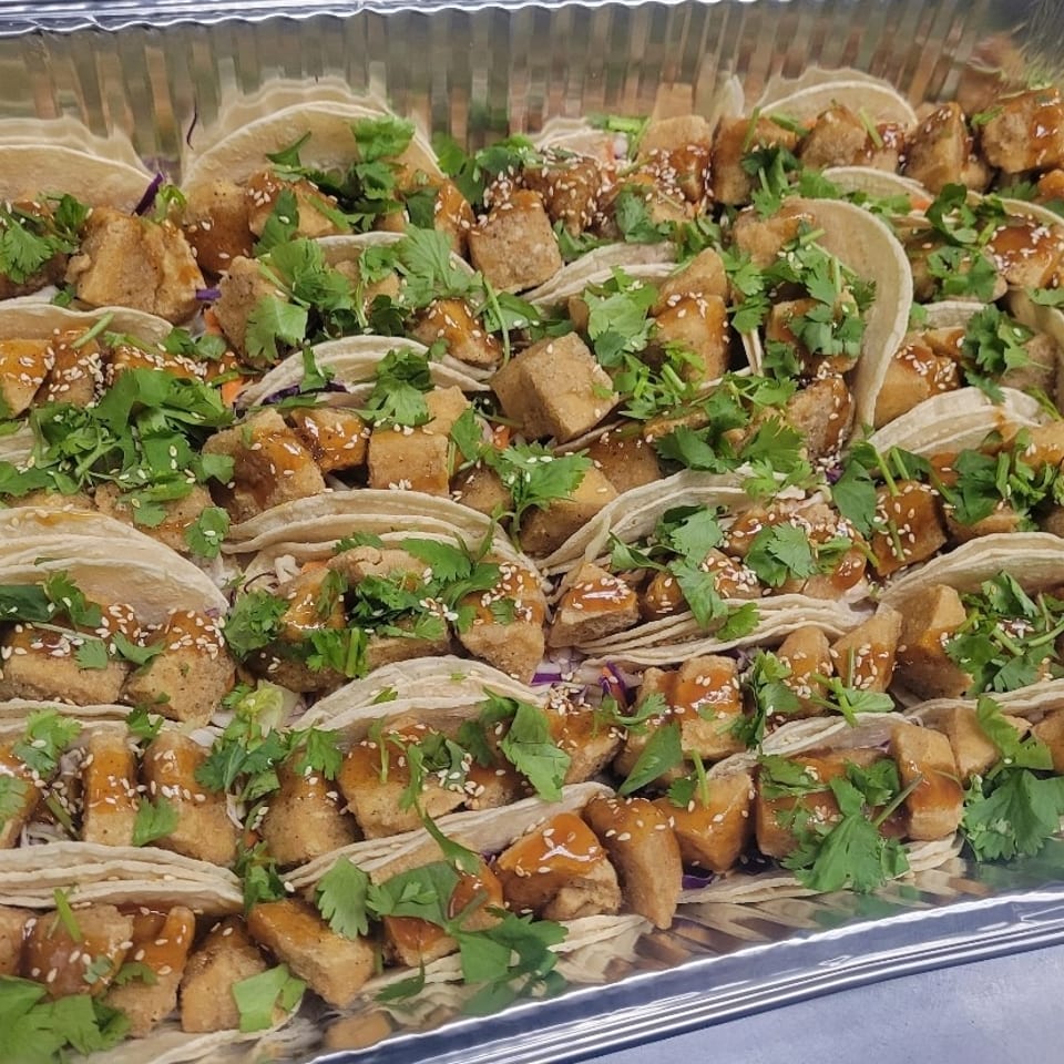 Tray of soft tacos filled with saucy crispy tofu, shredded cabbage, cilantro, and sesame seeds.