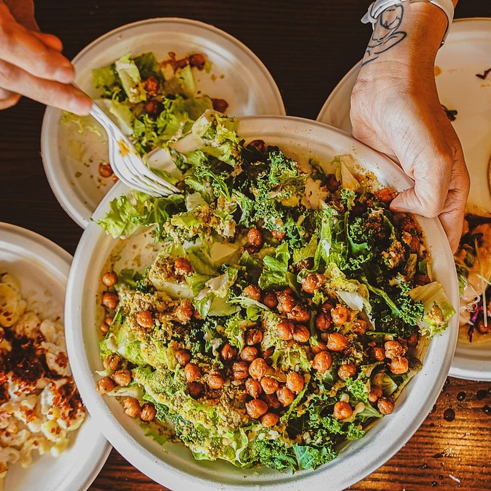 Hands serve a leafy green salad with roasted chickpeas alongside loaded fries, pasta, and grilled food on a wooden table.