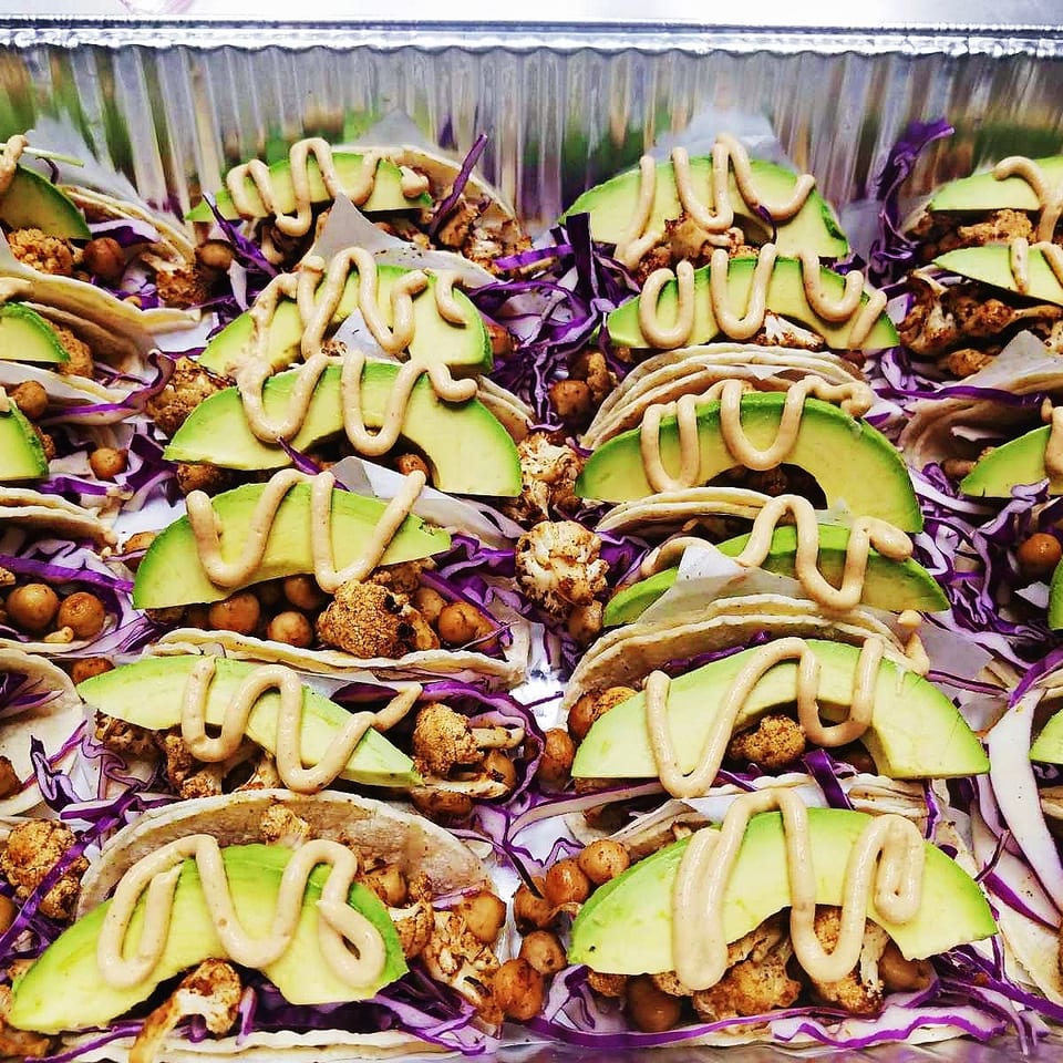 Tray of tacos filled with roasted cauliflower, chickpeas, purple cabbage, avocado slices, and drizzled creamy sauce.