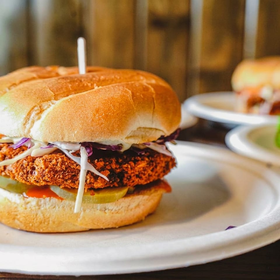 Crispy fried chicken sandwich with coleslaw and pickles on a plate, with additional sandwiches blurred in the background.
